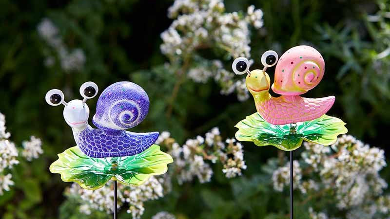 Two colorful snail garden stakes with a blurred green and white plant background.