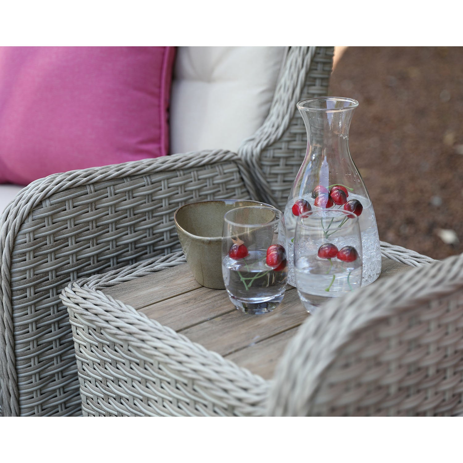 Outdoor setting with wicker furniture, a glass of water with cherries, and a carafe on a wooden table.