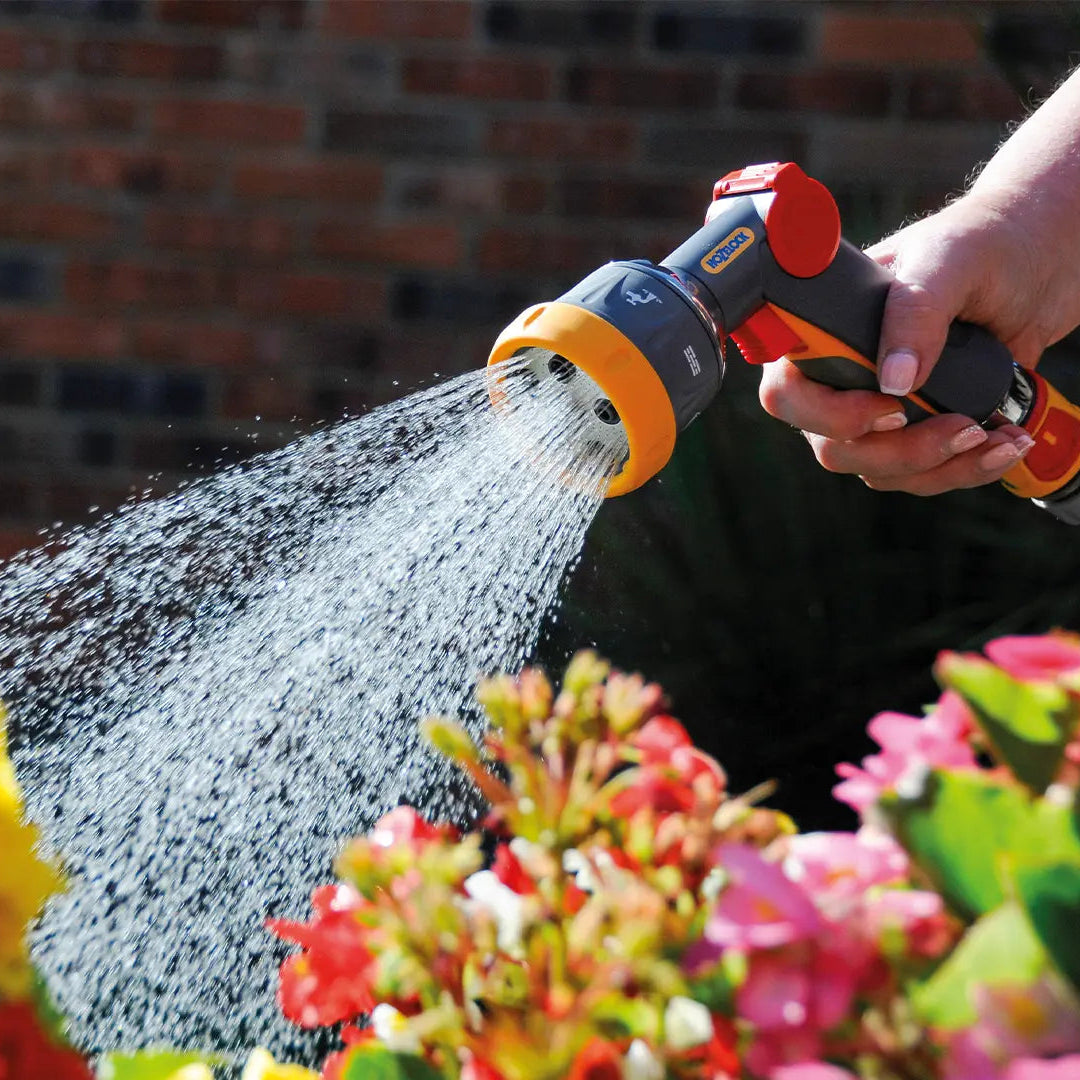 Person watering flowers with a garden hose nozzle against a brick wall.