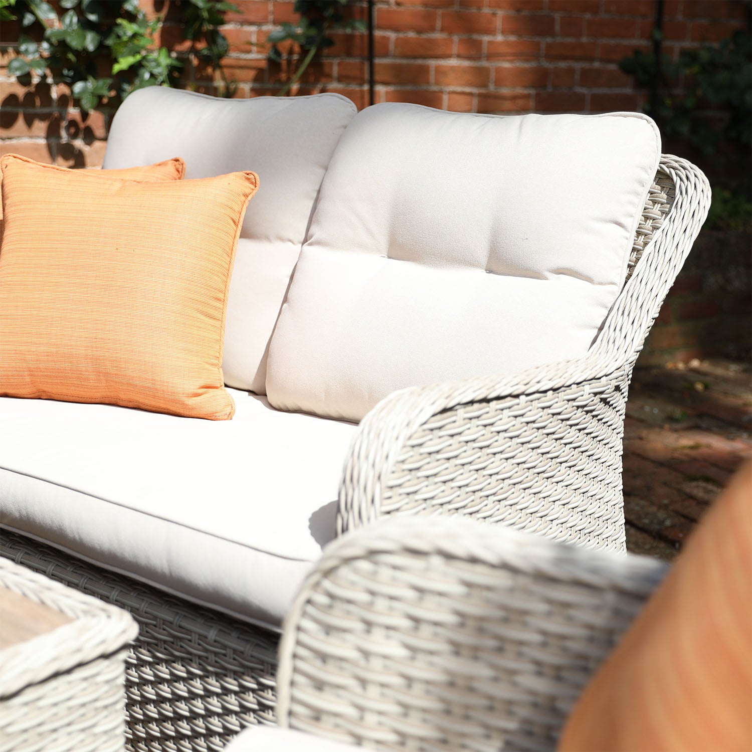 Wicker outdoor furniture with white cushions and an orange pillow against a brick wall.